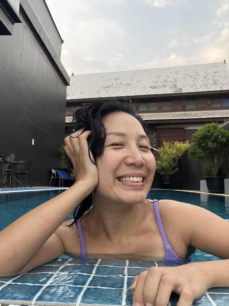 Falls leaning by the edge of a swimming pool, smiling at the camera.