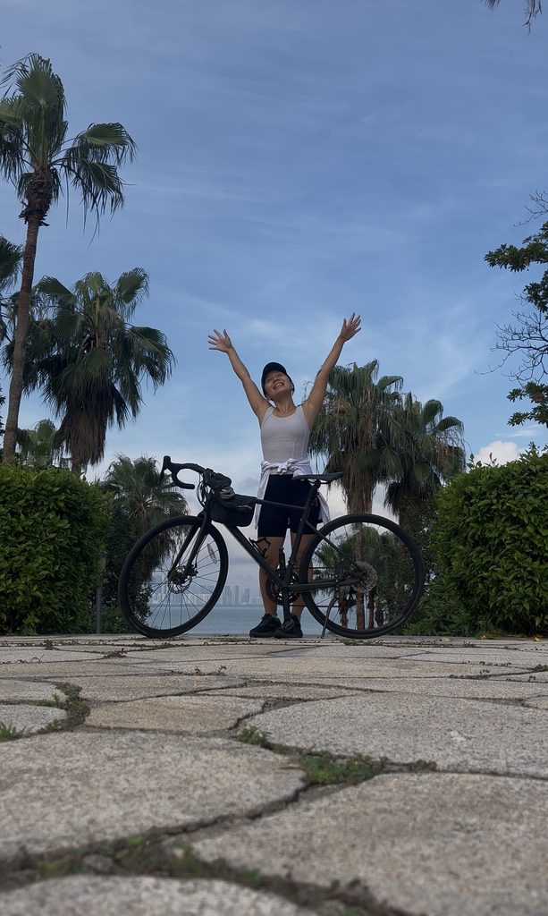 Falls standing by her bike at the seaside, raising both hands toward the sky and smiling with pure joy