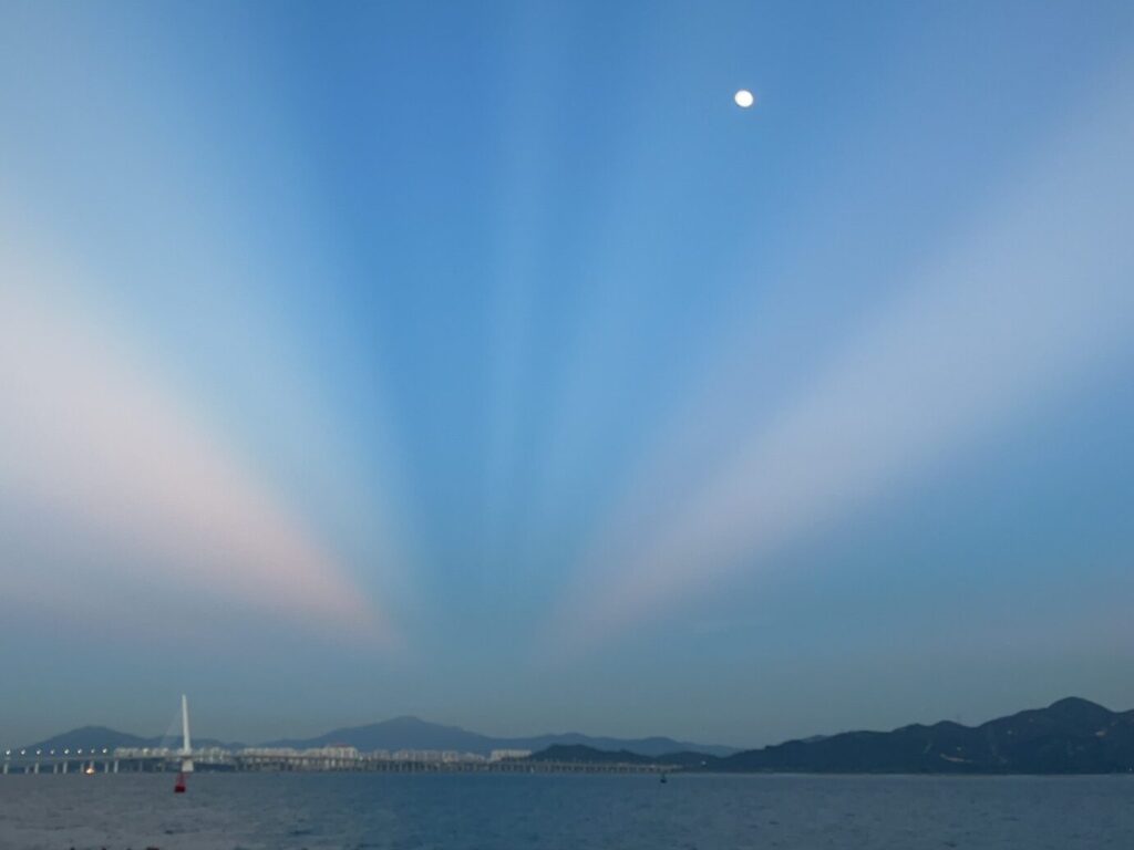 An evening scene with the moon already visible in the sky, overlooking the ocean and a glowing bridge in the distance
