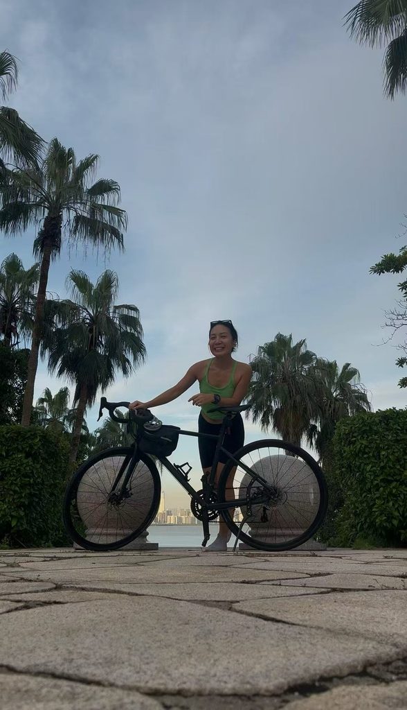 smiling at the camera while leaning against her bicycle on a seaside platform overlooking the ocean.