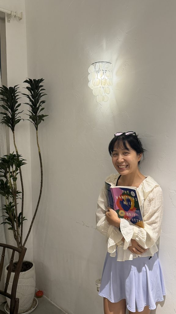 Falls holding The Vanishing Half book while posing beside a wall with a tree in the background