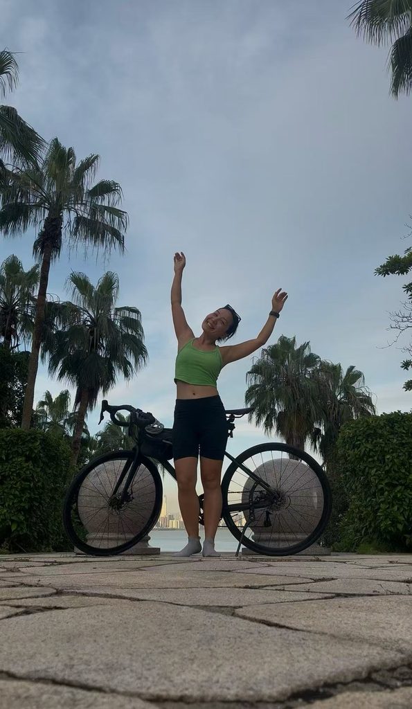 Falls smiling and raising her hand while leaning against her bicycle on a scenic coastal platform.