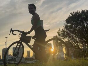 Falls walking across a grassy field at sunset while pushing her bicycle