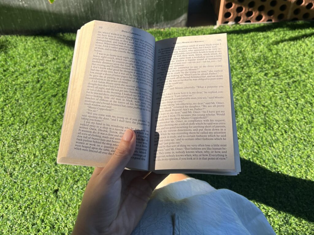 Holding an open book outdoors in the sunlight, surrounded by greenery.